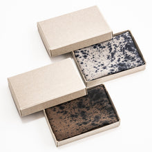 Three rectangular open boxes with beige lids, each containing a square cosmetic bar with a mottled speckled pattern. Two bars are dark brown/black and one is light gray/black, arranged on a clean white background.