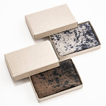Three rectangular open boxes with beige lids, each containing a square cosmetic bar with a mottled speckled pattern. Two bars are dark brown/black and one is light gray/black, arranged on a clean white background.