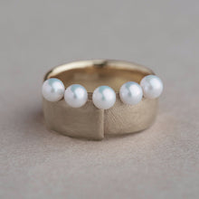 A gold ring featuring five small white pearls set along the front, photographed on a neutral textured beige background.