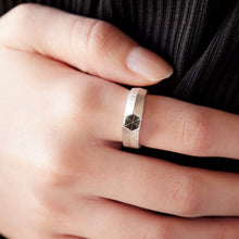 A close-up of a hand wearing a minimalist silver band ring with a faceted geometric star detail on the side, set against a dark ribbed garment.