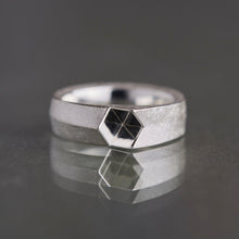 A silver geometric ring with a faceted hexagonal center set with a black diamond, displayed on a reflective dark gray surface.