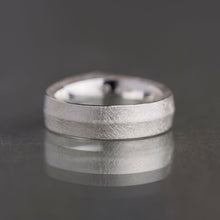 A textured silver band ring with a polished beveled edge, resting on a dark gray reflective surface.