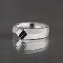 A silver geometric ring with a textured band and a faceted diamond-shaped inlay, resting on a dark reflective surface.