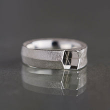 A silver geometric ring with a textured band and a faceted triangular side detail, resting on a dark reflective surface.