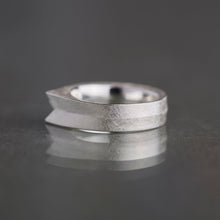 A textured silver ring with a unique tapered, stepped design, resting on a dark reflective surface against a dark gray background.