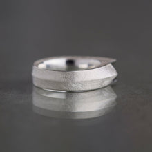 A textured silver band ring with a unique tapered, stepped design, resting on a dark reflective surface.