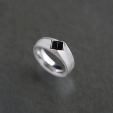 A silver band ring with a small black square inlay on the front, photographed on a dark gray textured background.