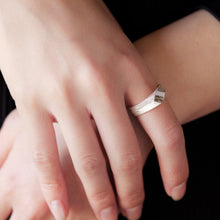A close-up of a person's hands wearing a minimalist silver band ring with a small geometric accent on one finger, set against a dark ribbed background.