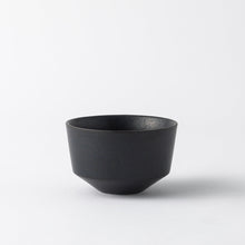 A minimalist black ceramic bowl with a textured matte finish and a geometric, tapered design, set against a clean white background.