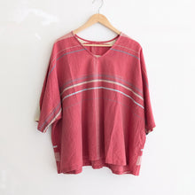A loose-fitting, short-sleeve V-neck top in dusty rose pink with thin horizontal stripes in beige, light blue, and white, hanging on a wooden hanger against a white wall.