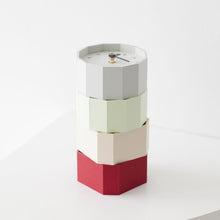 A modern stacked geometric wall clock with a hexagonal design, featuring four color-blocked tiers in gray, pale green, cream, and red, with a minimalist white face on top.